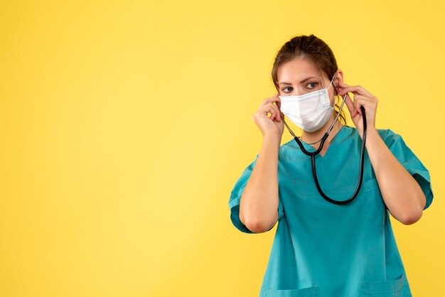 Medico femminile di vista frontale in camicia medica e maschera con lo stetoscopio su fondo giallo