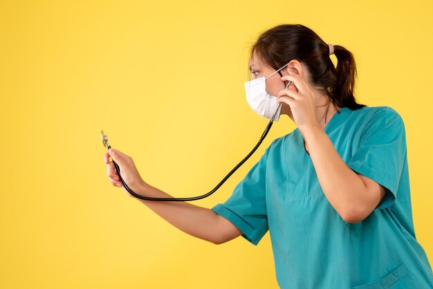 Medico femminile di vista frontale in camicia medica e maschera con lo stetoscopio su fondo giallo