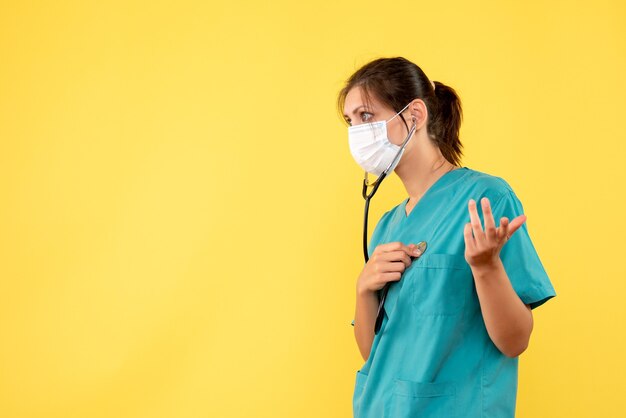 Medico femminile di vista frontale in camicia medica e maschera con lo stetoscopio su fondo giallo