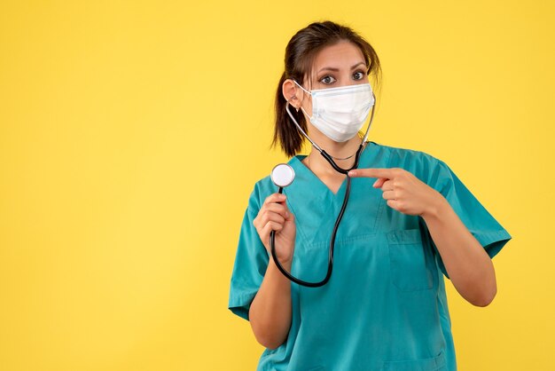 Medico femminile di vista frontale in camicia medica e maschera con lo stetoscopio su fondo giallo