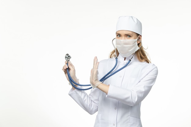 Medico femminile di vista frontale in vestito medico bianco con maschera sterile a causa del coronavirus che utilizza uno stetoscopio sulla malattia del muro bianco covid- malattia pandemica