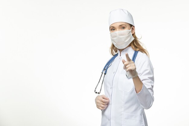 Medico femminile di vista frontale in vestito medico bianco con maschera sterile a causa di coronavirus sulla malattia pandemica del muro bianco covid