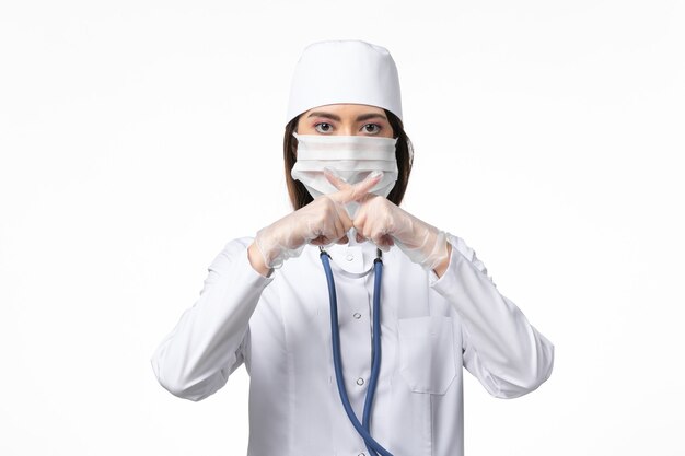 Medico femminile di vista frontale in vestito medico bianco con una maschera a causa della pandemia sul virus della malattia della malattia della parete bianca covid pandemico