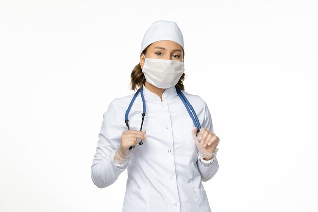 Medico femminile di vista frontale in vestito medico bianco e maschera sulla medicina di malattia malattia virus pandemia muro bianco chiaro