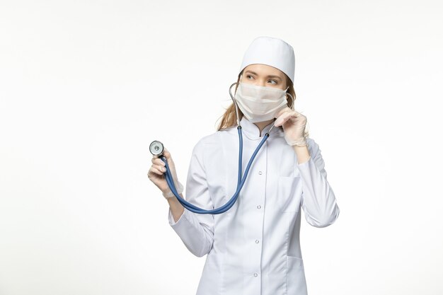 Medico femminile di vista frontale in vestito medico che indossa la maschera a causa dello stetoscopio della tenuta del coronavirus sulla malattia covid pandemica della parete bianca