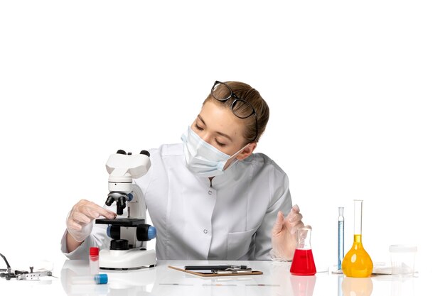 Medico femminile di vista frontale in vestito medico che indossa la maschera a causa di covid utilizzando il microscopio su uno spazio bianco chiaro