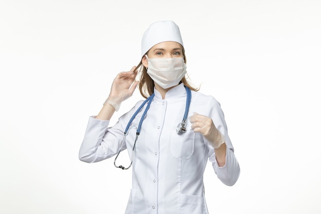 Medico femminile di vista frontale in vestito medico che indossa maschera a causa di coronavirus sulla malattia covid virus pandemico muro bianco