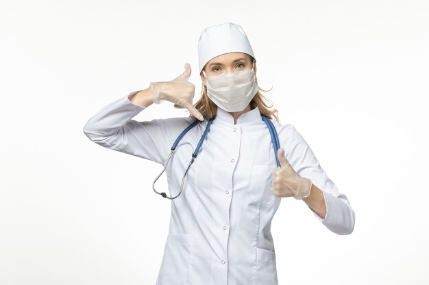 Medico femminile di vista frontale in vestito medico che indossa maschera e guanti a causa del coronavirus sul virus della malattia covid pandemica della scrivania bianca chiara