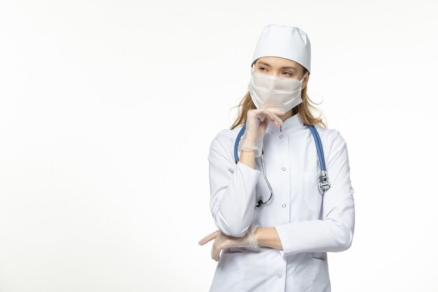 Medico femminile di vista frontale in vestito medico che indossa maschera e guanti a causa del pensiero del coronavirus sulla malattia da virus della malattia di covid pandemica del muro bianco