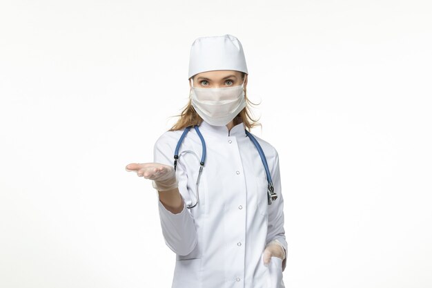 Medico femminile di vista frontale in vestito medico che indossa maschera e guanti a causa di coronavirus sulla scrivania bianca covid- malattia da virus della malattia pandemica