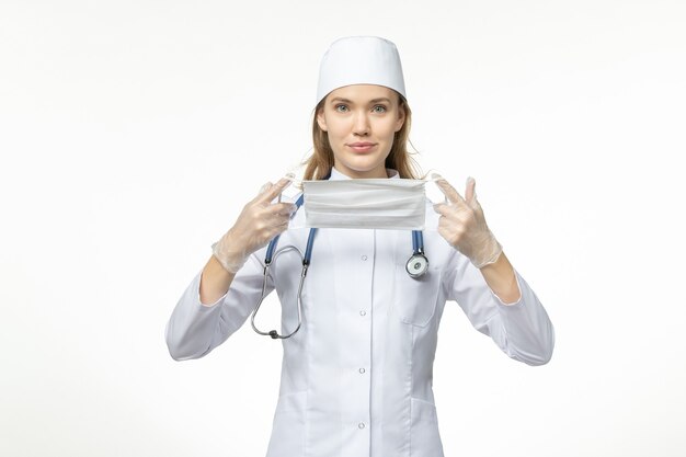 Medico femminile di vista frontale in vestito medico che tiene maschera a causa di coronavirus sulla malattia covid virus pandemico muro bianco