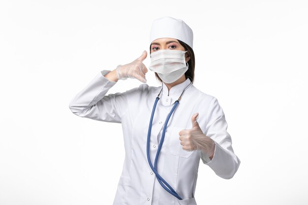 Medico femminile di vista frontale in vestito medico sterile bianco con maschera a causa del coronavirus sul virus covid della malattia pandemica del muro bianco