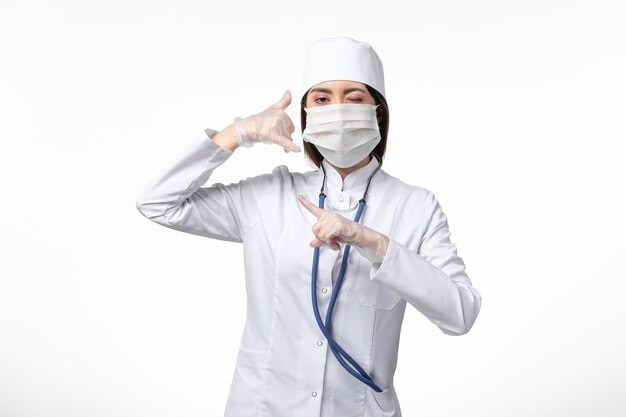 Medico femminile di vista frontale in vestito medico sterile bianco con maschera a causa di coronavirus su malattia pandemica di malattia del muro bianco chiaro covid- virus