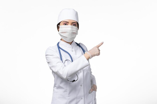 Medico femminile di vista frontale in vestito medico sterile bianco e maschera da portare a causa di covid- in posa sul muro bianco chiaro malattia virus covid- malattia pandemica