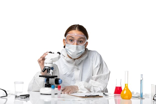 Medico femminile di vista frontale in vestito protettivo e con la maschera facendo uso del microscopio su fondo bianco coronavirus pandemico del virus di covid di salute