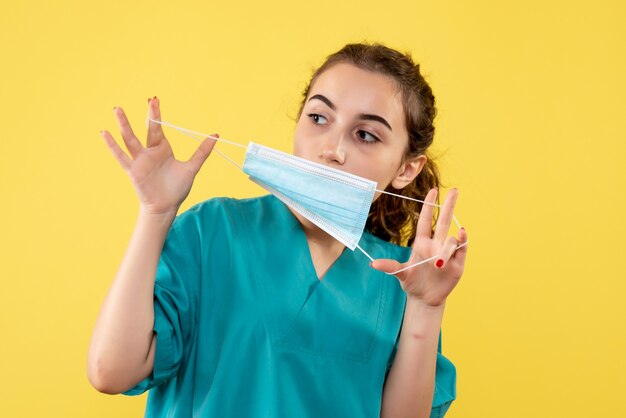 Medico femminile di vista frontale nella mascherina medica della tenuta della camicia sulla salute di covid di emozione di colore uniforme del virus della scrivania gialla