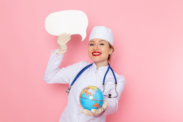 Medico femminile giovane vista frontale in vestito medico bianco con lo stetoscopio blu che tiene globo e segno bianco che sorride sulla salute medica dell'ospedale medico della medicina dello spazio rosa