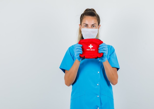 Medico femminile in uniforme blu, maschera, guanti che tengono il kit di pronto soccorso e che osserva attento