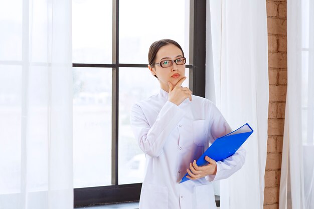 Medico femminile in vetri che tengono cartella blu vicino alla finestra.