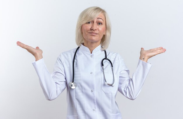 Medico femminile slava adulto confuso in abito medico con stetoscopio che tiene le mani aperte isolate su fondo bianco con lo spazio della copia