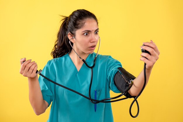 Medico femminile stupito vista frontale in uniforme utilizzando sfigmomanometri in piedi su sfondo giallo