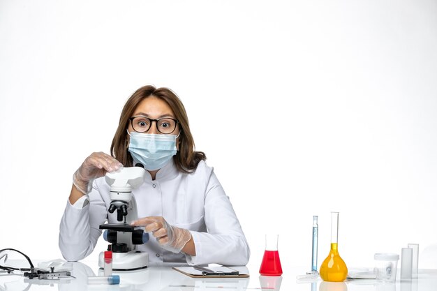 Medico femminile vista frontale in tuta medica bianca e maschera a causa di covid utilizzando il microscopio su uno spazio bianco chiaro