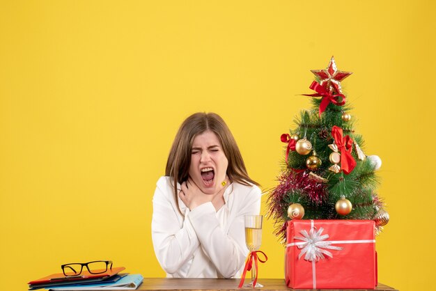 Medico femminile vista frontale seduto davanti al suo tavolo con mal di gola su sfondo giallo con albero di Natale e scatole regalo