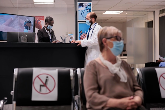 Medico generico in chat con l'addetto alla reception dell'ospedale durante la pandemia di coronavirus. Medico seduto nella sala d'attesa dell'atrio della struttura e parlando con il lavoratore del servizio sanitario.