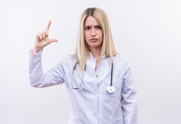 medico giovane ragazza bionda che indossa uno stetoscopio e abito medico che mostra le dimensioni sul muro bianco isolato