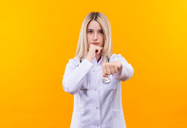 medico giovane ragazza che indossa stetoscopio in abito medico in piedi nella posa di combattimento sulla parete gialla isolata