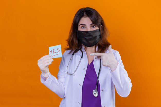 Medico invecchiato centrale indossando camice bianco in maschera protettiva nera e con stetoscopio tenendo la carta promemoria con sì parola che punta a esso