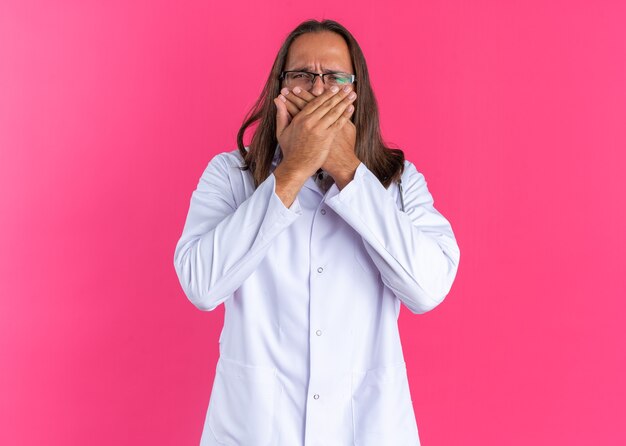 Medico maschio adulto accigliato che indossa tunica medica e stetoscopio con gli occhiali che tiene le mani sulla bocca guardando la telecamera isolata sul muro rosa