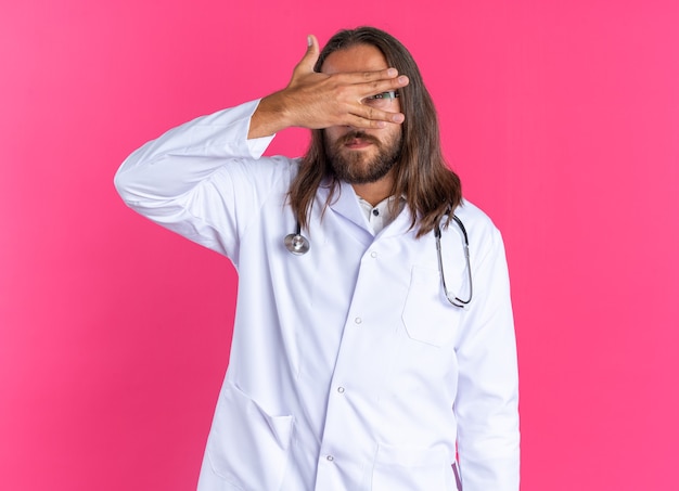 Medico maschio adulto spaventato che indossa accappatoio medico e stetoscopio con occhiali tenendo la mano davanti agli occhi guardando la telecamera tra le dita isolate sul muro rosa