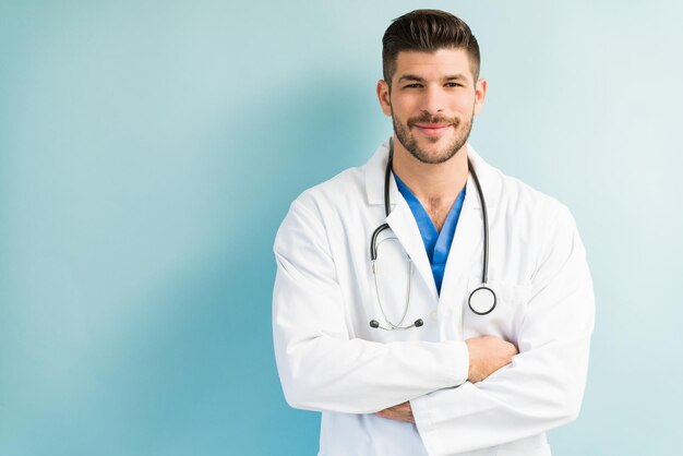 Medico maschio attraente sicuro che indossa un camice bianco da laboratorio mentre è in piedi con le braccia incrociate su sfondo turchese