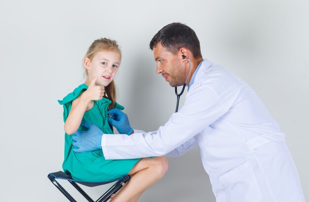 Medico maschio che ascolta il battito cardiaco mentre il bambino mostra il pollice in su in uniforme bianca e sembra allegro