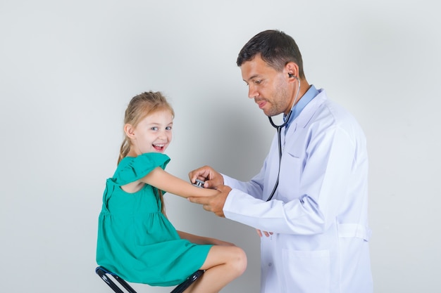 Medico maschio che esamina il braccio della bambina con lo stetoscopio nella vista frontale uniforme bianca.