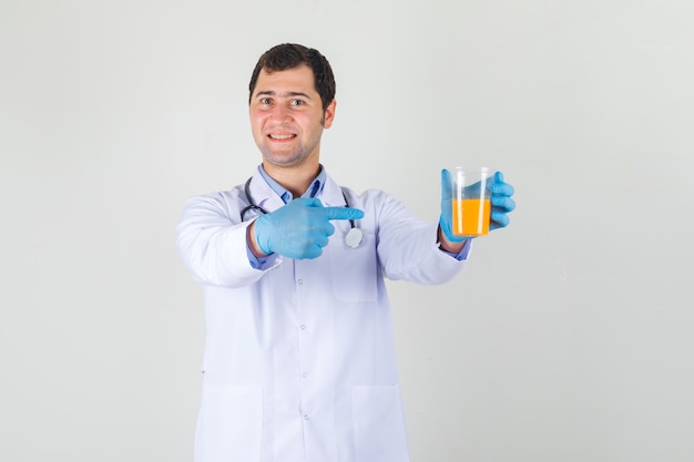 Medico maschio che indica il dito al bicchiere di succo in camice bianco, guanti e sembra allegro