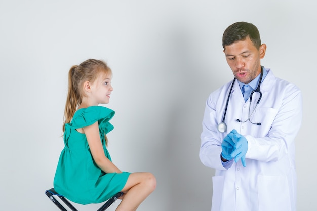 Medico maschio che indossa guanti in camice bianco con bambina e guardando attento. vista frontale.