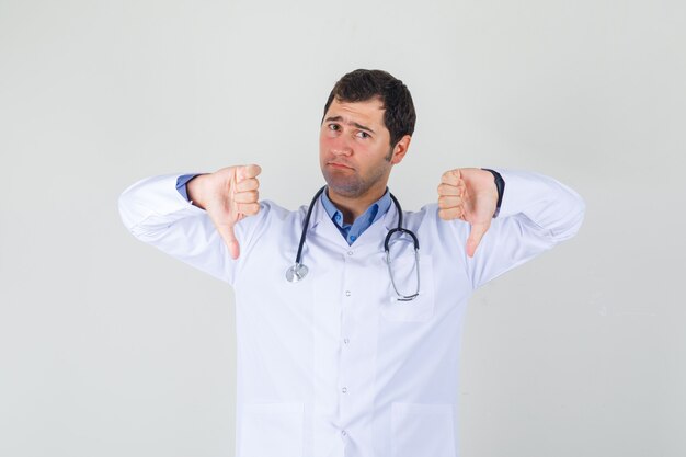 Medico maschio che mostra i pollici giù in camice bianco e sembra deluso. vista frontale.