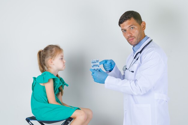 Medico maschio che mostra le pillole mentre il bambino si siede esaurito nella vista frontale uniforme bianca.