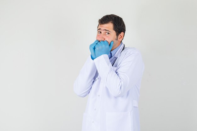 Medico maschio che tiene i pugni sulla bocca in camice bianco, guanti e sembra spaventata