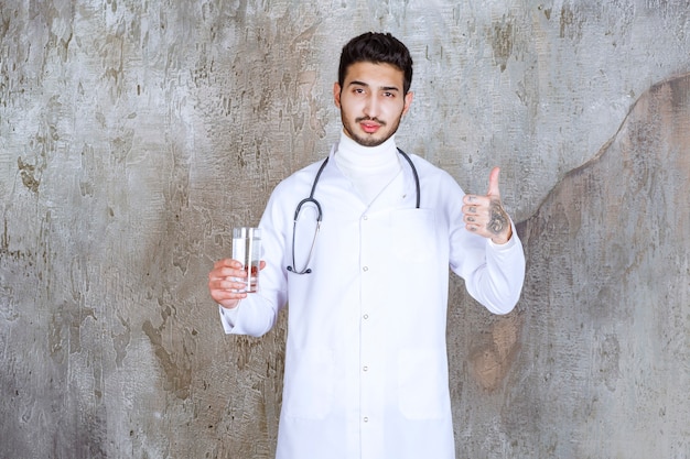 Medico maschio con lo stetoscopio che tiene un bicchiere di acqua pura e che mostra il segno positivo della mano.