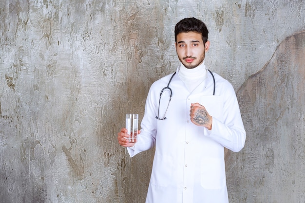 Medico maschio con lo stetoscopio che tiene un bicchiere di acqua pura e che mostra il segno positivo della mano