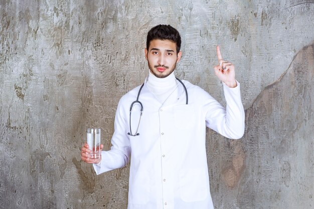 Medico maschio con lo stetoscopio che tiene un bicchiere di acqua pura e pensa a qualcosa.