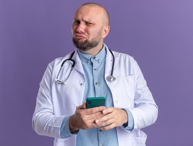 Medico maschio di mezza età confuso che indossa abito medico e stetoscopio che tiene il telefono cellulare con le labbra increspate isolate sulla parete viola