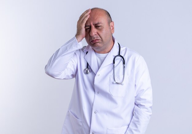 medico maschio di mezza età dolorante che indossa abito medico e stetoscopio tenendo la mano sulla testa guardando verso il basso isolato sul muro bianco
