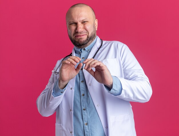 Medico maschio di mezza età irritato che indossa abito medico e stetoscopio che fa gesto di rifiuto isolato sulla parete rosa