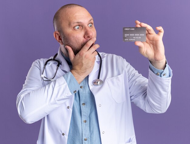 Medico maschio di mezza età preoccupato che indossa abito medico e stetoscopio che tiene e guarda la carta di credito tenendo la mano sulla bocca isolata sul muro viola