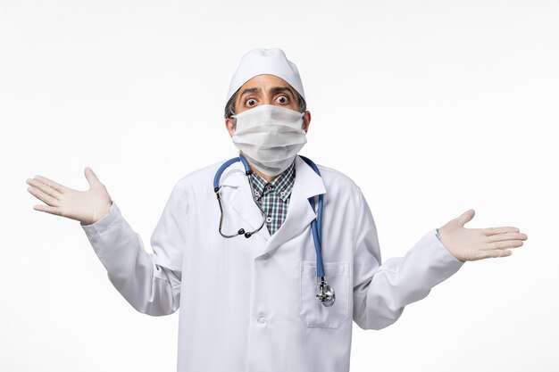 Medico maschio di vista frontale in tuta medica bianca a causa del coronavirus che indossa la maschera sulla superficie bianca