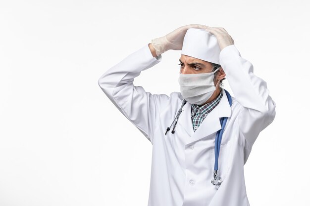 Medico maschio di vista frontale in tuta medica bianca con maschera a causa del pensiero del coronavirus sulla superficie bianca